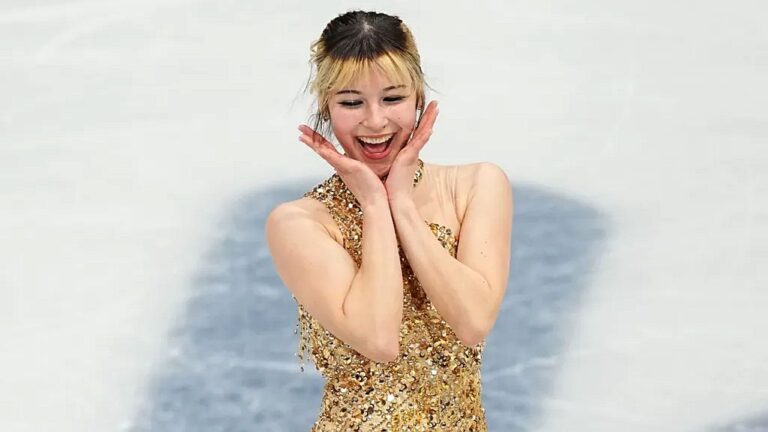 Patinadora artística sorri com expressão de surpresa e alegria, levando as mãos ao rosto enquanto se apresenta no gelo. Ela veste figurino dourado com detalhes brilhantes, em contraste com a pista branca ao fundo, sugerindo momento de comemoração após a performance.