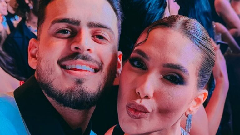 Homem e mulher tiram selfie em evento noturno com iluminação colorida ao fundo. Ele sorri para a câmera, enquanto ela faz biquinho, usando maquiagem marcante e brincos longos. Outras pessoas aparecem desfocadas ao redor, sugerindo ambiente de festa ou premiação.