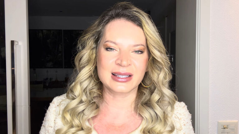 Joice Hasselmann de cabelos loiros ondulados, brincos de argola dourados e casaco claro, olhando para a câmera e falando em ambiente interno iluminado