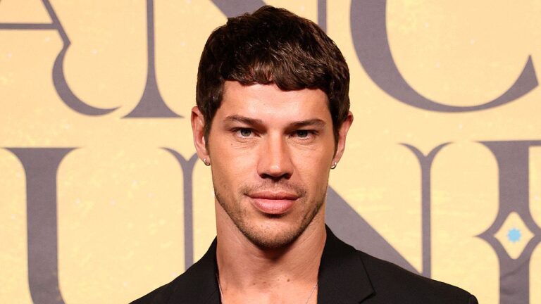 Ator José Loreto encara a câmera vestindo paletó preto, posicionado à frente de um painel bege com letras cinzas grandes e desfocadas ao fundo