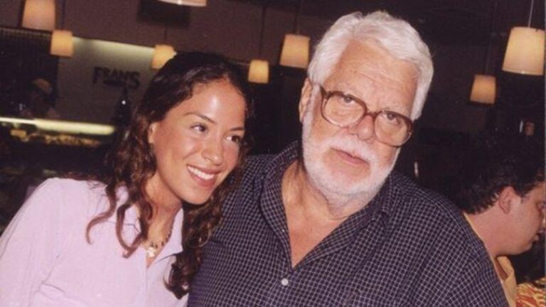 Homem idoso de cabelos e barba brancos, usando óculos e camisa escura, posa ao lado de mulher sorridente em ambiente interno com iluminação aconchegante, sugerindo encontro social ou familiar.