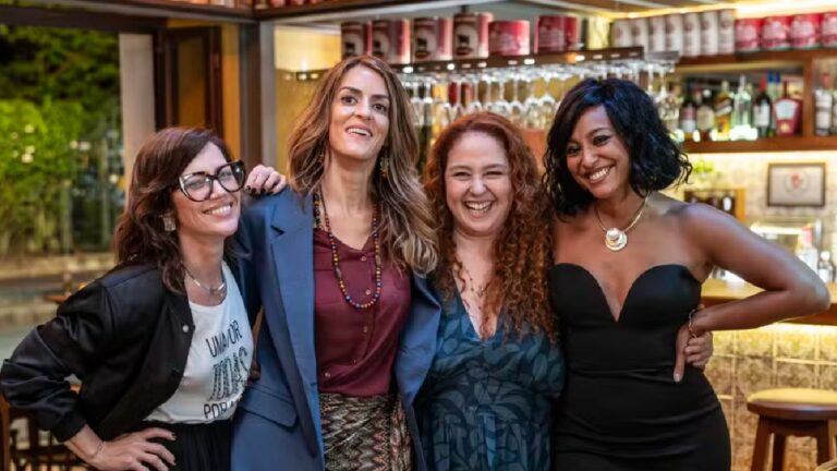 Quatro mulheres posam abraçadas e sorridentes em ambiente de bar ou restaurante, com prateleiras de bebidas iluminadas ao fundo, em clima descontraído e de confraternização.