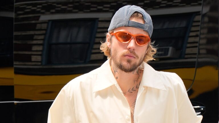 Homem jovem com barba e cabelo loiro escuro na altura dos ombros usa boné cinza virado para trás e óculos de sol com armação laranja. Ele veste camisa clara de gola aberta, exibindo tatuagens no pescoço, e está ao ar livre diante de um veículo escuro ao fundo, com expressão séria e postura descontraída.