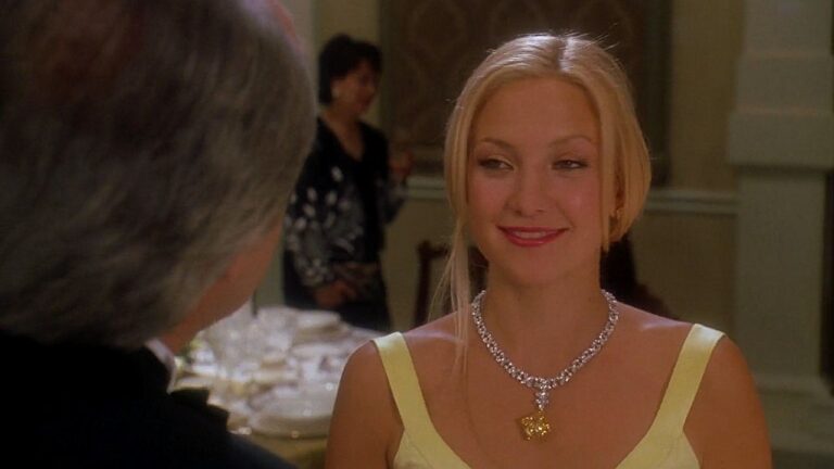 Mulher loira usa vestido claro e colar de diamantes durante jantar formal. Ela sorri suavemente para a pessoa à sua frente, em salão elegante com mesa posta ao fundo e iluminação quente, sugerindo evento social sofisticado.