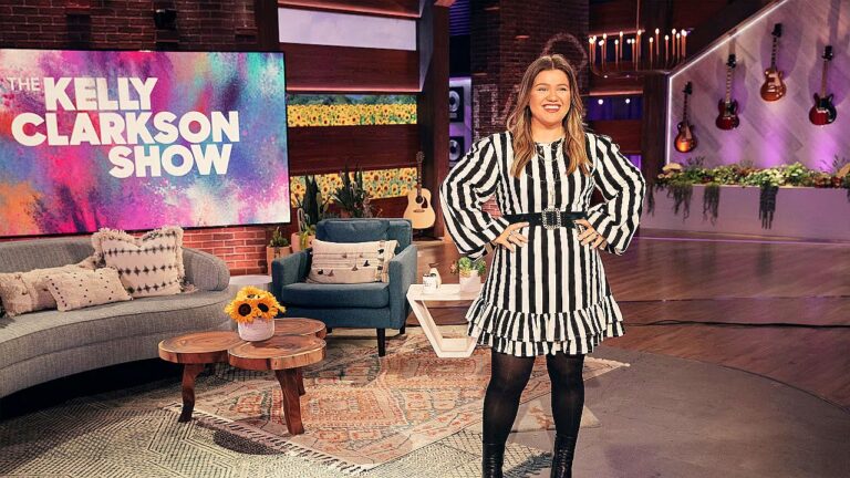 Apresentadora posa no palco do programa “The Kelly Clarkson Show”, sorrindo com as mãos na cintura. Ela veste vestido listrado em preto e branco com cinto largo, meia-calça escura e botas. Ao fundo, o estúdio decorado inclui sofá, poltronas, mesas de centro com flores, guitarras na parede e um grande telão com o nome do programa.