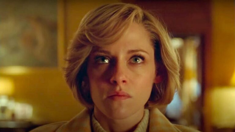 Mulher de pele clara em close frontal ocupa o centro da imagem, com cabelo loiro curto cortado na altura do queixo e levemente ondulado para o lado direito, olhos claros bem abertos e fixos à frente transmitindo expressão séria e tensa, sobrancelhas levemente franzidas e lábios fechados sem sorriso, usando blusa de gola alta clara e casaco bege ou creme, em ambiente interno com iluminação quente e amarelada