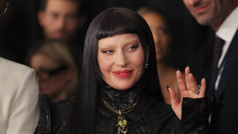 Pessoa com cabelo preto liso e franja curta sorri e acena, vestindo roupa preta de couro com colar dourado, em um evento com outras pessoas ao fundo.