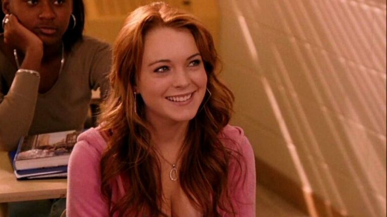 Jovem mulher ruiva, com cabelo longo e ondulado, sorri enquanto está sentada em uma sala de aula. Ela usa blusa rosa e colar delicado, além de brincos grandes. Ao fundo, outra estudante aparece sentada com livros sobre a mesa. O ambiente tem iluminação quente, e a expressão da jovem é alegre e atenta, como se estivesse participando de uma conversa ou aula.