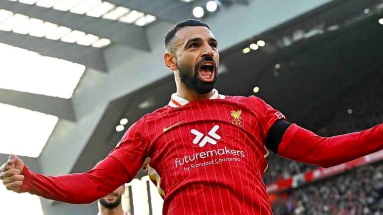Jogador de futebol com uniforme vermelho aparece em momento de comemoração intensa dentro do estádio. Ele está com os braços abertos e a boca aberta em grito, demonstrando euforia após um lance decisivo, enquanto torcedores e a estrutura do estádio aparecem ao fundo.