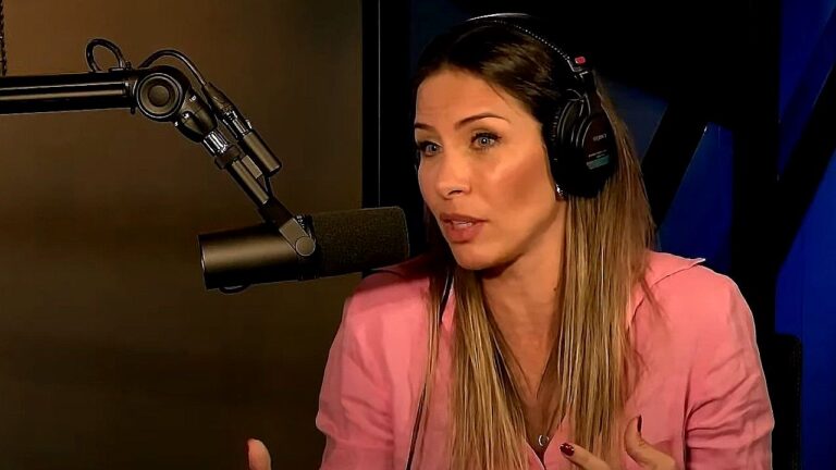 Mulher participa de entrevista em estúdio de podcast, usando fones de ouvido e falando ao microfone profissional montado em braço articulado. Vestindo blazer rosa, ela gesticula enquanto conversa, em ambiente fechado com iluminação controlada e fundo escuro.