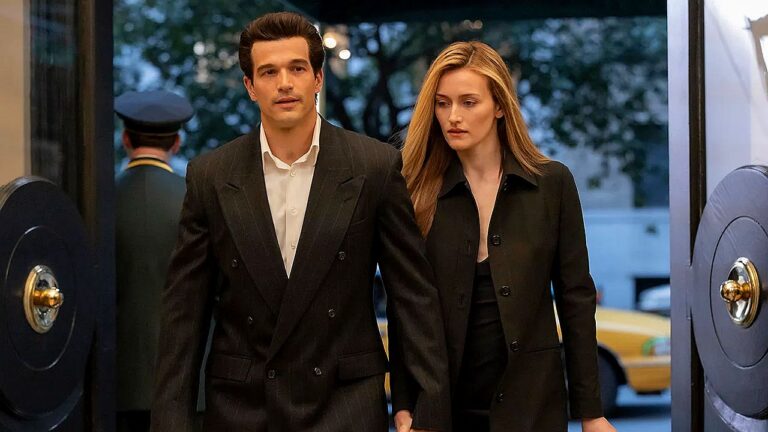 Um homem e uma mulher, ambos vestidos com roupas formais escuras, entram por uma porta elegante de vidro e metal. O homem usa terno preto com camisa branca e caminha à frente com expressão séria e focada. A mulher, também de preto, vem logo atrás, com semblante preocupado ou pensativo. Ao fundo, é possível ver um porteiro e um táxi amarelo na rua, sugerindo a entrada de um hotel ou prédio sofisticado no início da noite.