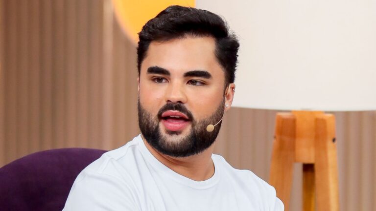 Lucas Guedez de camiseta branca, barba aparada e sobrancelhas marcadas, fala ao microfone de lapela em estúdio de televisão com fundo em tons claros