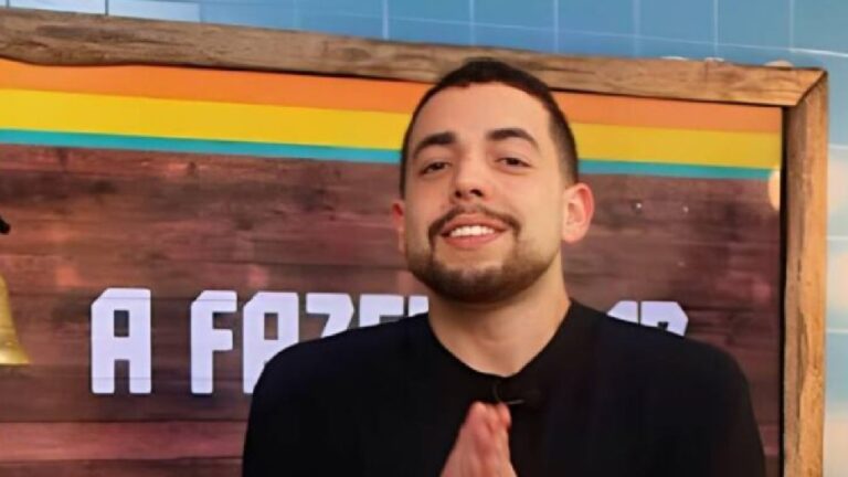 Homem jovem de barba e cabelo curto sorri para a câmera com as mãos juntas à frente do peito. Ele veste camiseta preta e está em um estúdio com painel de madeira ao fundo, onde se vê parte de um letreiro com letras brancas e faixa colorida na parte superior.