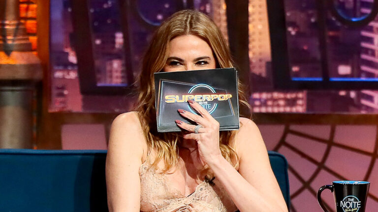 Apresentadora Luciana Gimenez sentada em sofá azul no programa The Noite, cobrindo parte do rosto com ficha do Superpop em cenário noturno de TV