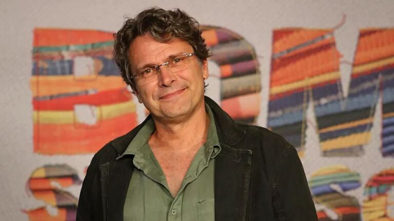 Homem de meia-idade posa sorridente diante de painel com letras coloridas ao fundo. Ele usa óculos, camisa verde aberta no colarinho e jaqueta escura, olhando diretamente para a câmera em ambiente que sugere evento cultural ou lançamento artístico.