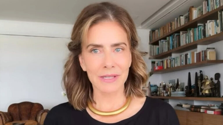 Maitê Proença de cabelos castanhos curtos, olhos claros e colar dourado rígido, vestindo blusa preta em frente a uma estante de livros e objetos diversos