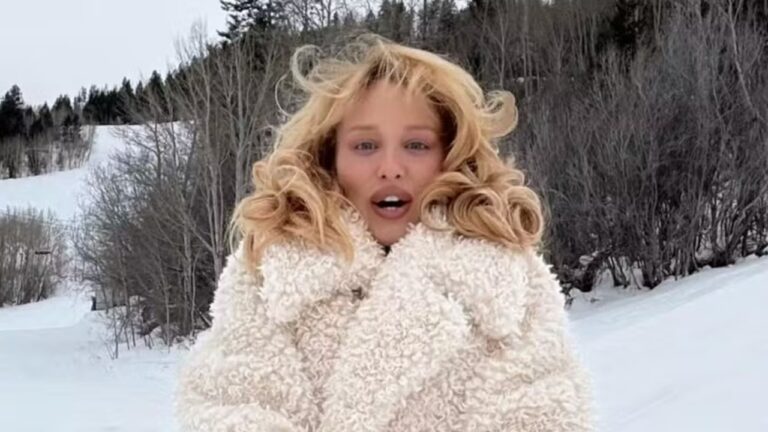 Mulher de casaco felpudo claro posa ao ar livre em cenário de neve e árvores ao fundo.