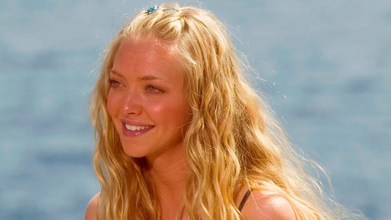 Mulher jovem de pele clara aparece em close ao ar livre com o mar azul claro desfocado ao fundo, cabelos longos loiros ondulados soltos com pequena mecha presa no topo da cabeça por acessório discreto, rosto levemente inclinado para a esquerda da imagem, olhos claros voltados para fora do enquadramento, sorriso suave mostrando os dentes, expressão tranquila e luminosa, pele com brilho natural sob luz solar direta