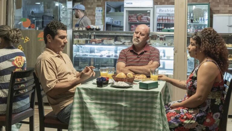 Três pessoas sentadas à mesa conversam em lanchonete com vitrine de salgados ao fundo em cena de filme brasileiro