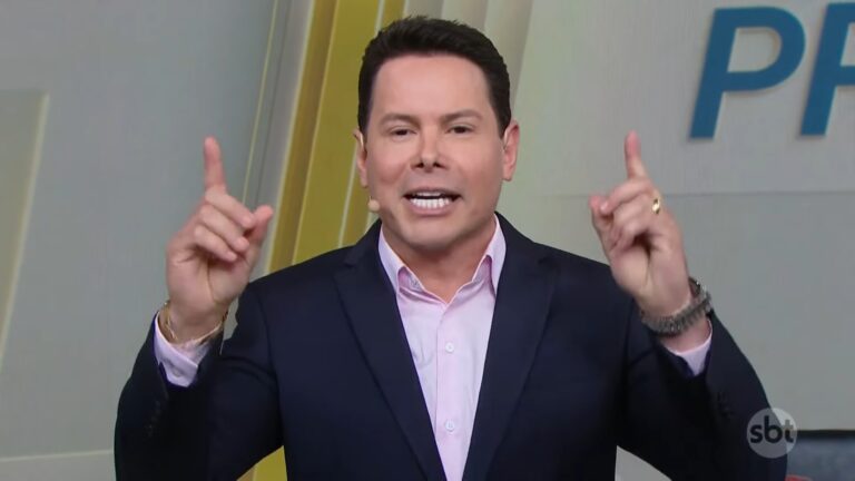 Marcão do Povo, de terno escuro e camisa rosa, aparece no estúdio do Primeiro Impacto, do SBT, gesticulando com as mãos erguidas enquanto fala. Ele usa microfone de lapela e fundo colorido com letras parciais de um logotipo, indicando que apresenta um programa ao vivo