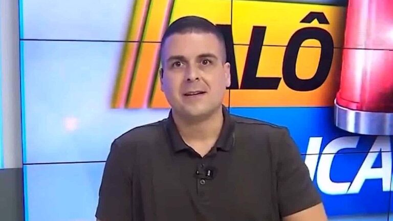 Homem aparece em estúdio de TV diante de um telão com gráficos coloridos e parte de uma palavra em destaque ao fundo. Ele usa camisa polo escura e microfone preso à gola, olhando diretamente para a câmera com expressão atenta, como se estivesse apresentando ou comentando uma notícia ao vivo.
