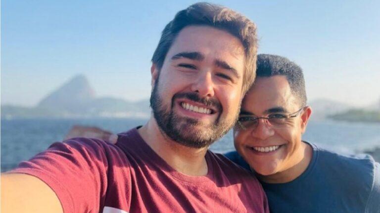 Dois homens aparecem sorrindo em uma selfie ao ar livre, com o mar ao fundo. Um deles está segurando a câmera com o braço estendido e usa camiseta vermelha; tem barba e cabelo castanho. O outro, ao lado, usa camiseta azul e óculos, com o braço apoiado no ombro do amigo. Ao fundo, é possível ver a água e uma paisagem costeira com uma formação rochosa ou montanha ao longe, sob céu claro.