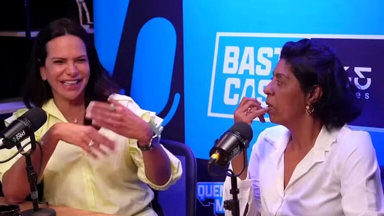 Duas mulheres participam de gravação em estúdio de podcast, sentadas diante de microfones profissionais. A mulher à esquerda, vestindo camisa clara, gesticula enquanto fala; a da direita, de blazer branco, escuta atentamente com a mão próxima ao rosto. Ao fundo, painel azul com iluminação de estúdio e parte do nome do programa em destaque.