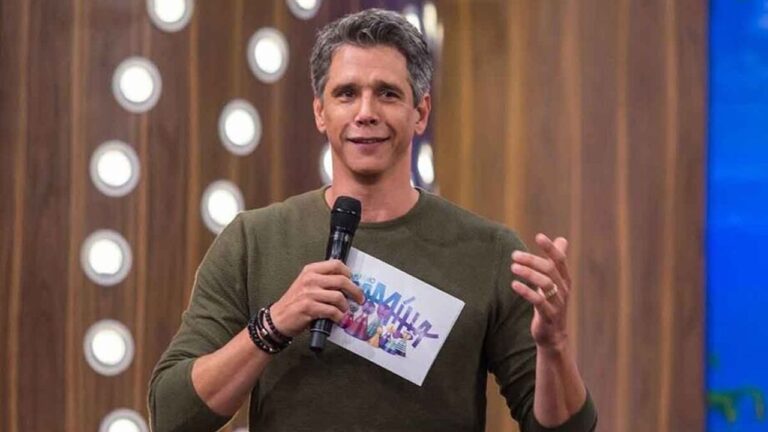 Homem de meia-idade com cabelos curtos grisalhos e pele clara aparece em estúdio de televisão, vestindo camiseta verde de mangas compridas, segurando um microfone preto com uma das mãos e um cartão branco com texto colorido na outra, usando pulseiras no pulso esquerdo e anel no dedo anelar, olhando para a câmera com leve sorriso e expressão confiante enquanto gesticula como se estivesse apresentando ou falando ao público, com fundo de cenário de programa de auditório composto por painéis de madeira e círculos luminosos brancos distribuídos verticalmente e iluminação de estúdio bem marcada