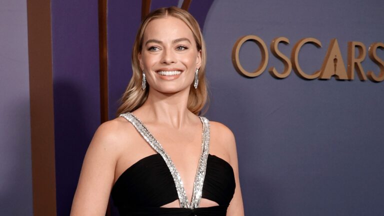 Margot Robbie sorri no tapete vermelho do Oscar usando vestido preto com detalhes prateados e brincos longos brilhantes