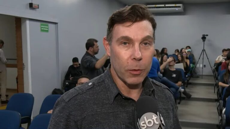 Maurício Abravanel na frente de um microfone logotipo do SBT enquanto é entrevistado em um auditório com várias pessoas sentadas ao fundo