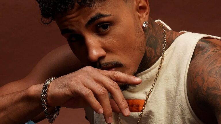 Homem posa em close, apoiando a mão no rosto e olhando diretamente para a câmera com expressão séria. Ele usa regata clara, corrente e pulseira douradas, brinco e exibe tatuagens no braço e no pescoço, em retrato com fundo liso em tom terroso.