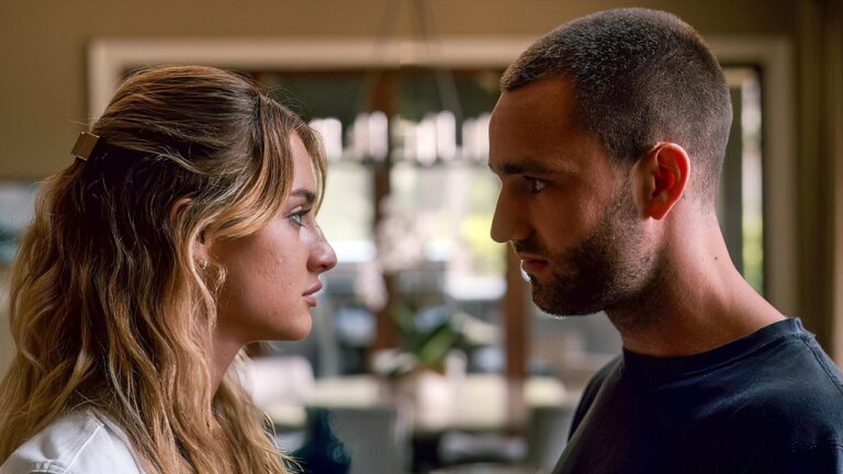 Um homem e uma mulher estão frente a frente, muito próximos, trocando um olhar intenso. Ela tem cabelos longos com mechas claras presos parcialmente atrás e veste uma blusa clara. Ele tem cabelo bem curto e barba aparada, usando uma camiseta escura. O ambiente parece ser interno, com luz natural entrando por janelas ao fundo. A expressão de ambos sugere uma conversa séria ou um momento emocionalmente carregado.