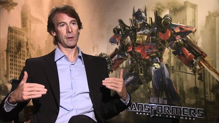 Diretor Michael Bay fala durante entrevista com pôster de Transformers ao fundo mostrando o robô Optimus Prime