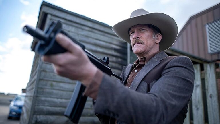 Homem com visual de faroeste aponta uma arma de fogo à frente. Ele usa chapéu de cowboy claro, blazer marrom e camisa escura, além de ter bigode. Está ao ar livre, diante de construções simples de madeira que lembram um cenário rural ou cidade pequena. A expressão dele é séria e determinada, sugerindo um momento de confronto ou tensão.
