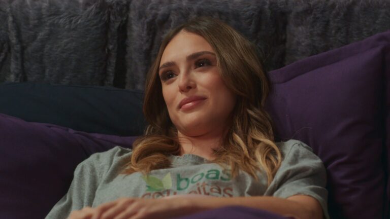 Naiane, interpretada por Isabelle Drummond, está deitada em uma cama com lençóis e travesseiros roxos e sorri levemente enquanto olha para frente. Ela veste uma camiseta cinza com estampa colorida e parece estar em um momento de tranquilidade em ambiente íntimo e acolhedor na novela Coração Acelerado