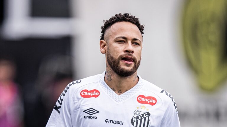 Homem atleta em campo de futebol visto da cintura para cima usa camisa branca de time com escudo preto no lado esquerdo do peito e logotipos de patrocinadores em vermelho e preto na parte frontal e nos ombros tem barba curta bem aparada cabelo curto com laterais raspadas e topo cacheado expressão concentrada com boca levemente aberta e língua visível olhando para frente possui tatuagens visíveis no pescoço e parte superior do peito o fundo está desfocado