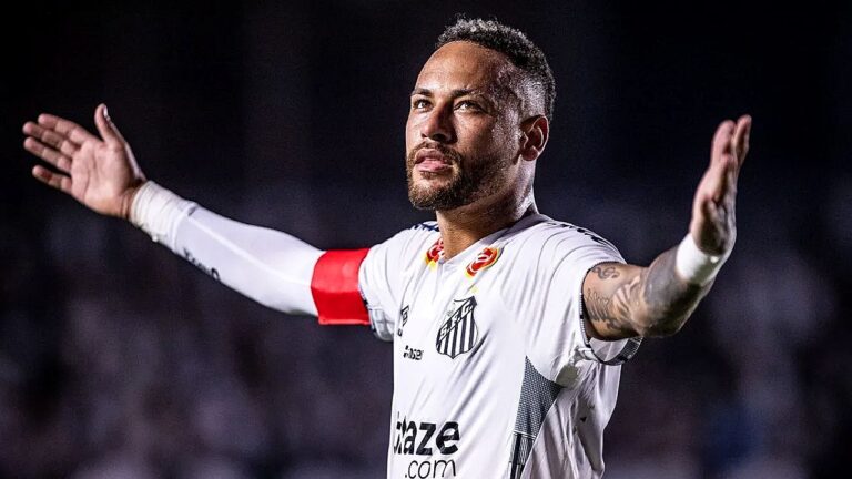 Jogador de futebol veste uniforme branco do Santos e usa braçadeira vermelha de capitão enquanto abre os braços em campo, sob iluminação de estádio. Com expressão intensa e olhar voltado para cima, ele parece comemorar ou reagir a um lance durante a partida, diante de arquibancadas desfocadas ao fundo.