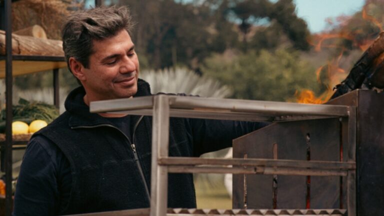 Felipe Bronze prepara churrasco em área externa enquanto manuseia a churrasqueira acesa durante gravação de programa culinário