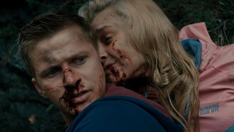 Homem e mulher aparecem feridos em ambiente externo de floresta, com marcas de sangue no rosto. O homem, em primeiro plano, olha para o lado com expressão tensa, enquanto a mulher, também machucada, se apoia nele por trás, aparentando cansaço ou dor. A cena sugere momento de perigo ou sobrevivência após situação violenta.