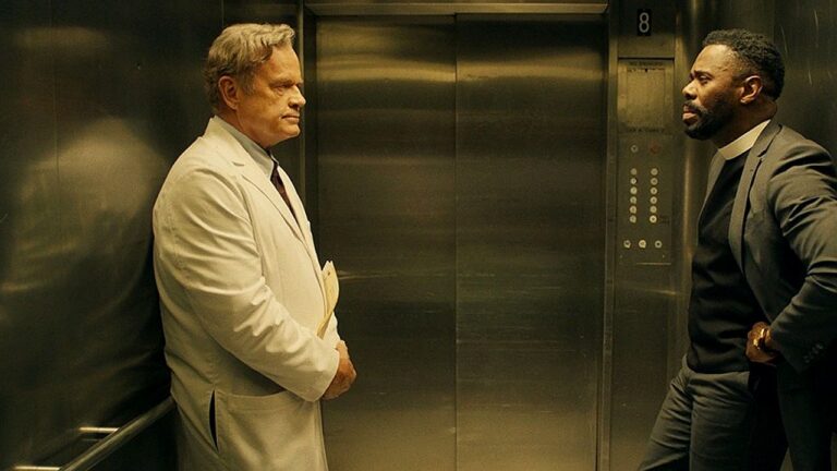 Dois homens estão dentro de um elevador com portas metálicas fechadas. À esquerda, um homem mais velho veste jaleco branco e segura uma pasta, enquanto encara o outro com expressão séria. À direita, um homem de terno escuro apoia uma das mãos na parede do elevador e parece falar de forma tensa, sugerindo confronto ou discussão em ambiente corporativo ou hospitalar.