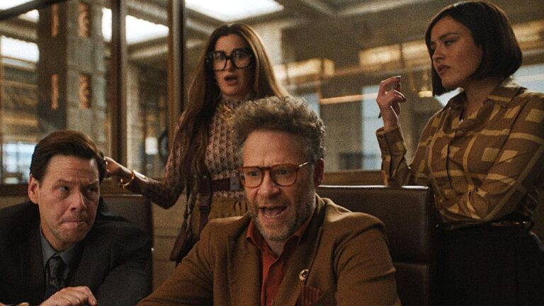 Quatro pessoas estão reunidas em um ambiente que parece ser um escritório ou sala de reuniões com paredes de vidro e iluminação quente. No centro, um homem de meia-idade com barba curta e óculos está sentado à mesa, vestindo blazer marrom e camisa escura. Ele parece estar falando com intensidade ou discutindo algo importante. À esquerda, outro homem, também de meia-idade, usa terno escuro e observa com expressão séria. Atrás, uma mulher de cabelo longo e óculos demonstra surpresa ou preocupação. À direita, outra mulher com cabelo curto, usando camisa estampada em tons terrosos, parece pensativa, levando a mão ao rosto.