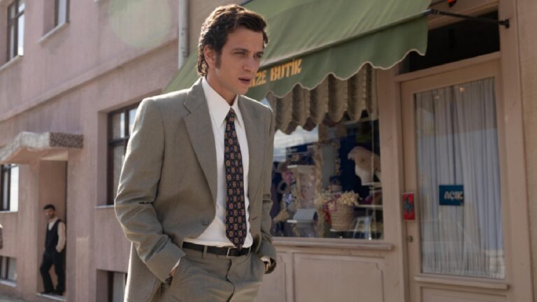 Homem jovem, de cabelo castanho ondulado, caminha pela calçada usando terno cinza e gravata estampada, com as mãos nos bolsos. Ele passa em frente a uma loja com toldo verde e vitrine decorada, em uma rua iluminada pela luz do dia.