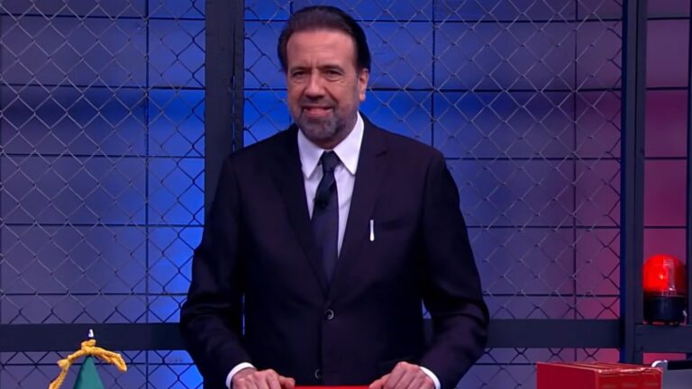 Jorge Lordello, de terno escuro e gravata, está no estúdio do Operação de Risco com fundo formado por grades metálicas e iluminação em tons de azul e vermelho. Ele está de pé, com as mãos apoiadas em uma bancada