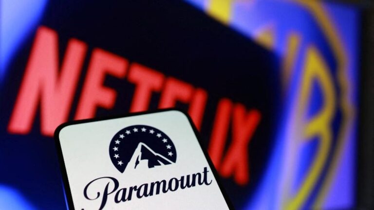 Imagem em close mostra um smartphone com o logotipo da Paramount+ em destaque na tela. Ao fundo, desfocado, aparece o nome “Netflix” em letras vermelhas e parte do logotipo da Blockbuster, sugerindo concorrência entre plataformas de streaming e marcas do setor de entretenimento.