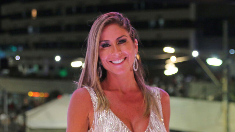 Patrícia Maldonado sorri em evento noturno, a jornalista usa vestido prateado com brilhos, brincos de argola cravejados e fundo com luzes de cidade e prédios