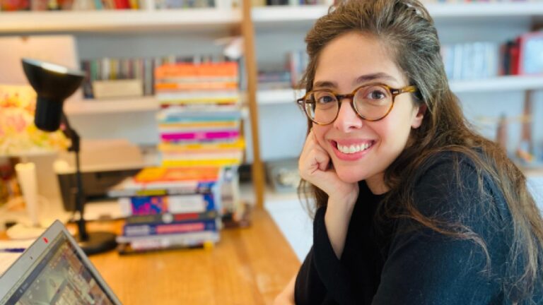 Mulher jovem com óculos de armação redonda sorri para a câmera enquanto está sentada a uma mesa de trabalho. Ela apoia o rosto na mão e usa blusa preta. À sua frente há um notebook aberto, e ao fundo aparecem estantes com livros organizados e uma luminária de mesa acesa, sugerindo ambiente de estudo ou home office iluminado e acolhedor.