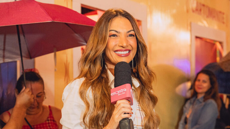 Patrícia Poeta sorri de batom vermelho segurando microfone do programa Encontro em ambiente externo com guarda-chuva bordô e pessoas ao fundo