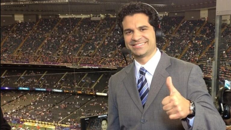 Homem aparece em primeiro plano dentro de um estádio esportivo coberto, usando terno cinza, camisa branca e gravata listrada em tons de azul, com fone de ouvido profissional e microfone acoplado à cabeça, sugerindo transmissão ao vivo.