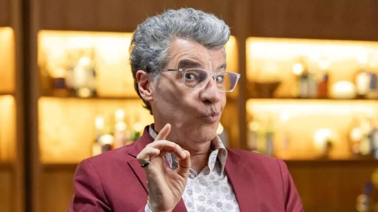 Homem de cabelos grisalhos e óculos de armação transparente faz expressão divertida, com os lábios contraídos e a mão levantada em gesto delicado, como se enfatizasse um ponto. Ele veste blazer vermelho sobre camisa clara estampada. Ao fundo, prateleiras iluminadas exibem garrafas, sugerindo um bar ou estúdio com cenário aconchegante.