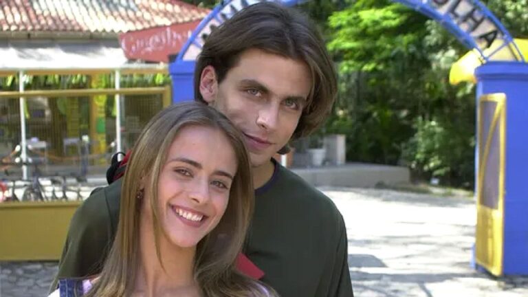 Um casal jovem posa abraçado ao ar livre, sorrindo para a câmera. A mulher apoia a cabeça no ombro do rapaz, enquanto ele a envolve com o braço. Ao fundo, há árvores, construções coloridas e um arco decorativo, sugerindo parque ou espaço público em dia ensolarado.